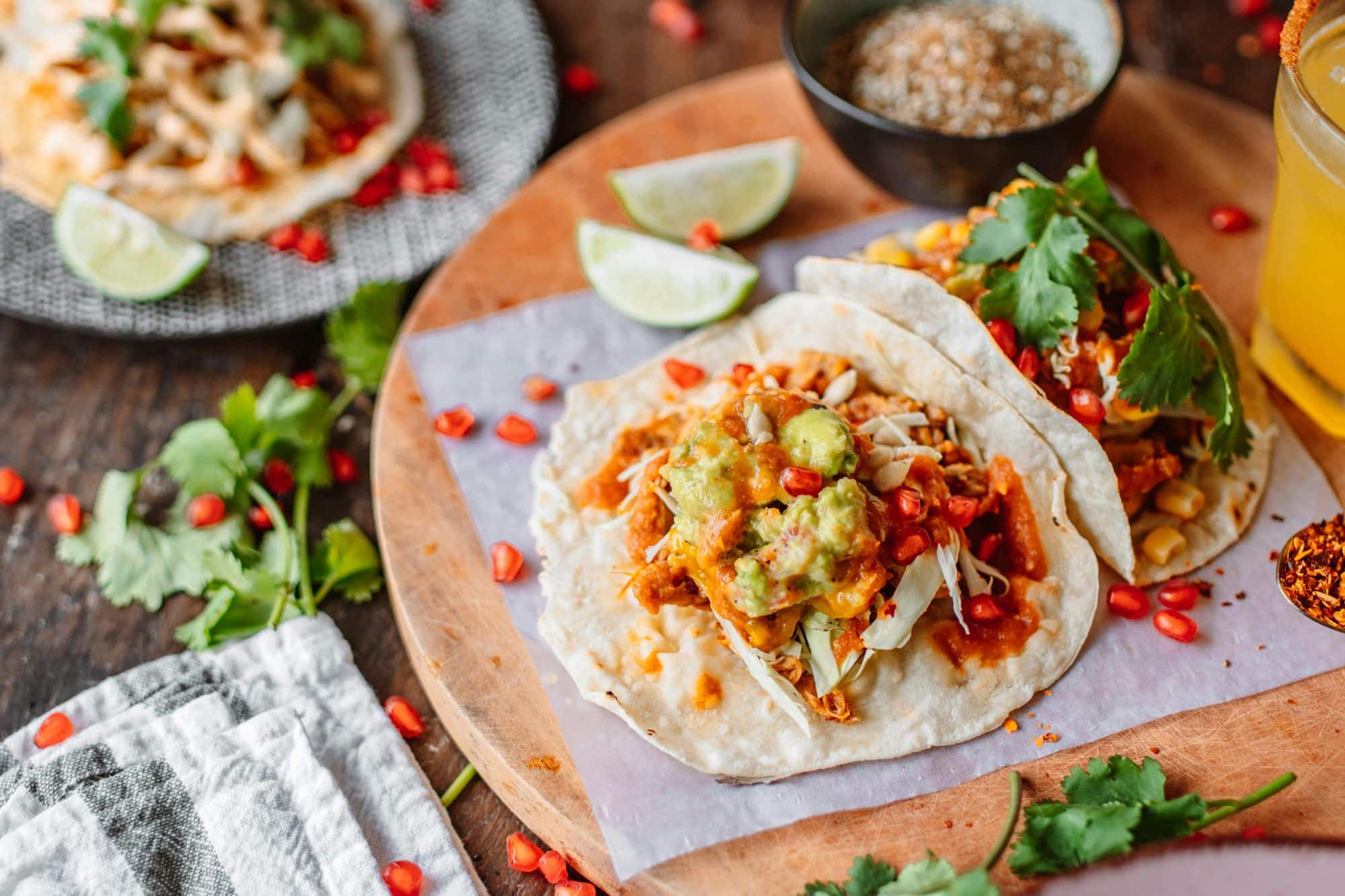Two tacos topped with guacamole, shredded veggies, and sauce on a wooden board, surrounded by limes and herbs in San Tan Valley, AZ