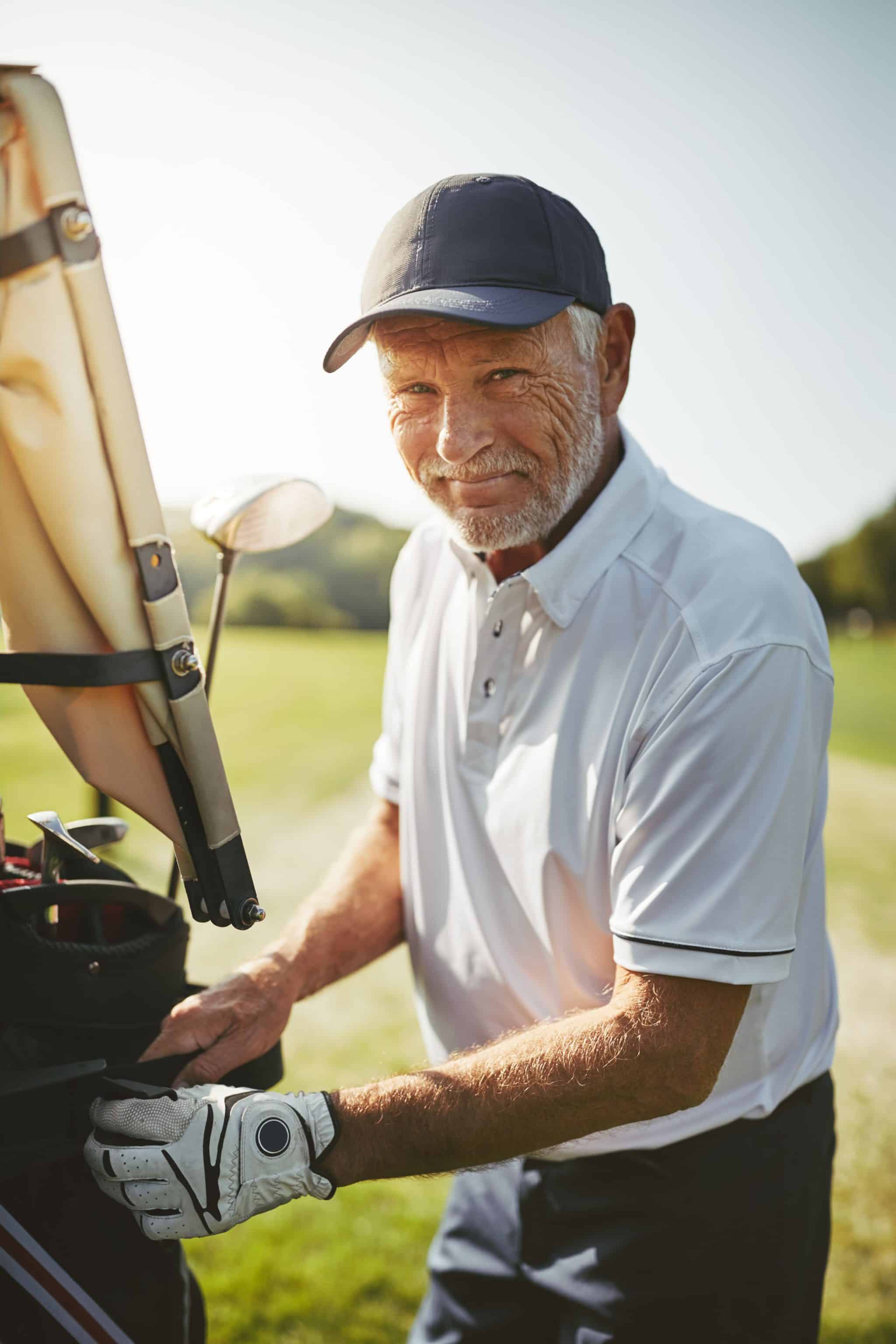 Older man in a white polo and cap smiling while standing next to a golf bag on a sunny course in San Tan Valley, AZ