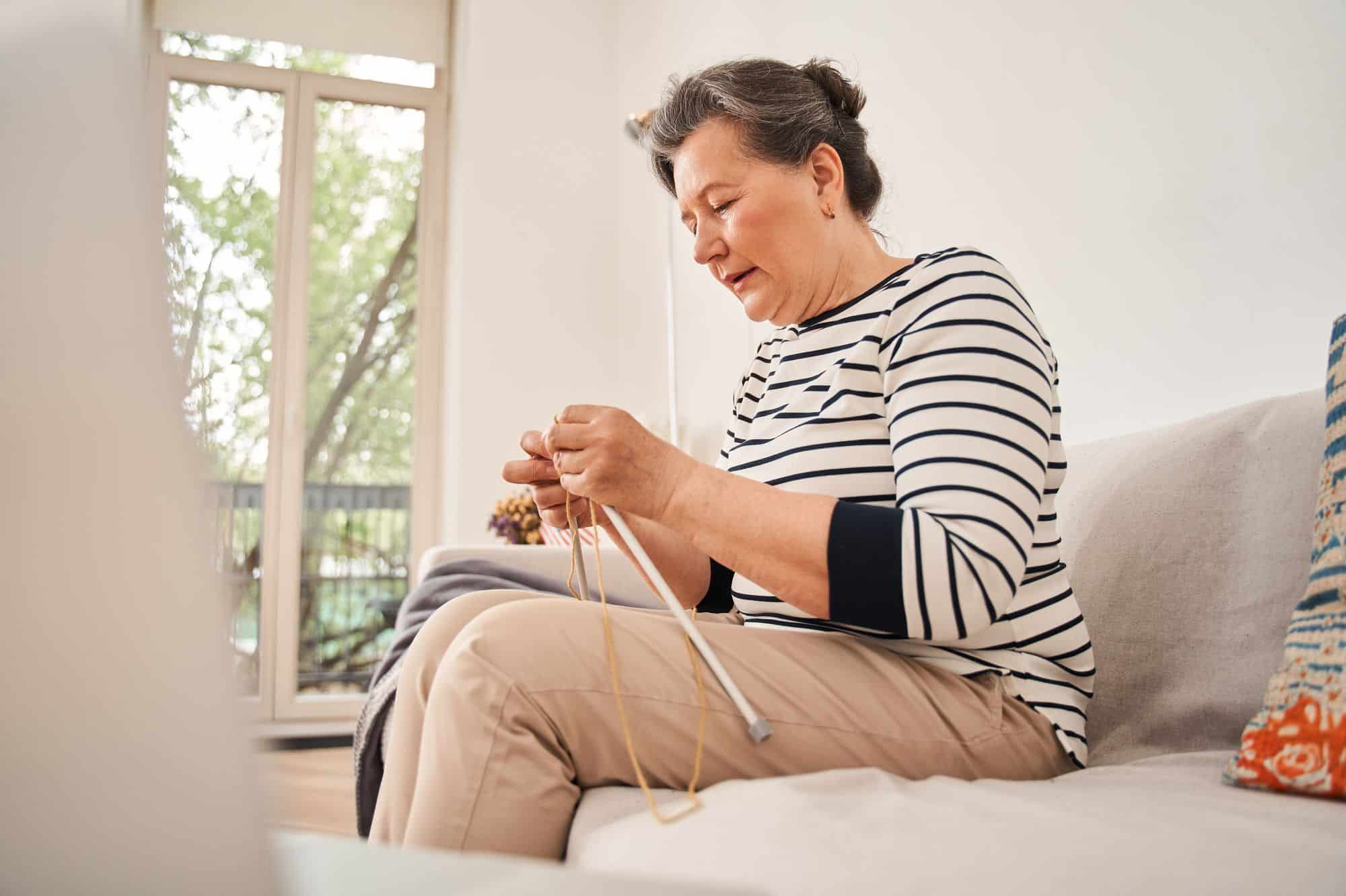 A woman sitting on a couch near a window, focused on knitting with light brown yarn and needles in San Tan Valley, AZ