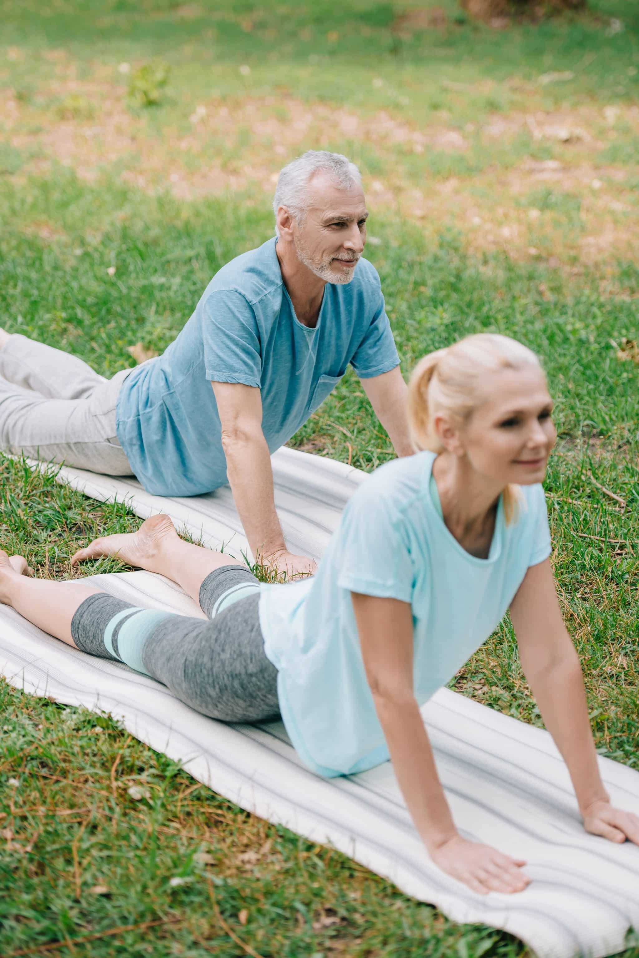 Older man and woman doing cobra pose on yoga mats outside on grass, with trees in the background in San Tan Valley, AZ